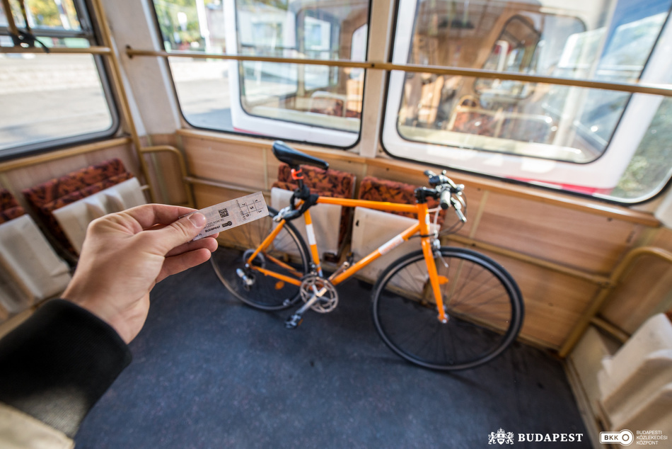 A villamos egyik utasa a menetjegyét mutatja a kamerának, a háttérben egy rögzített kerékpár látható